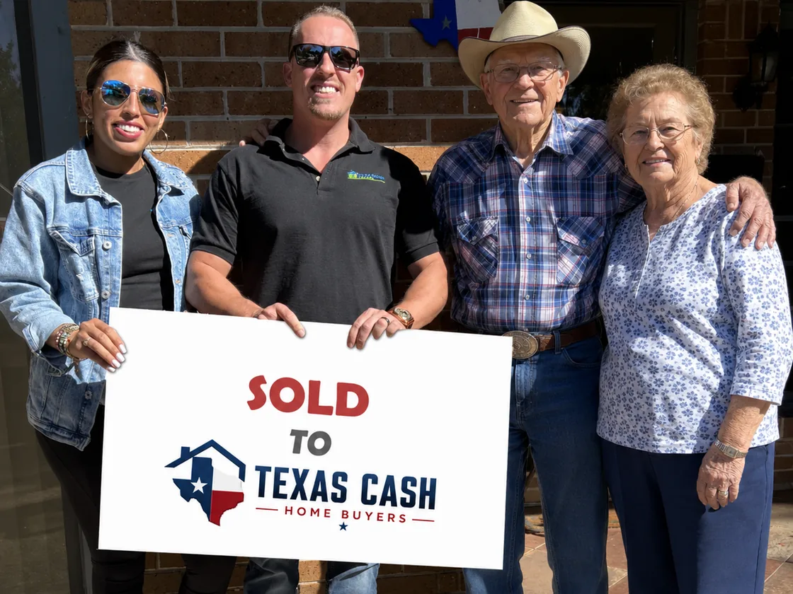 Four people standing together outside a brick house, holding a large sign reading Sold To Texas Cash Home Buyers